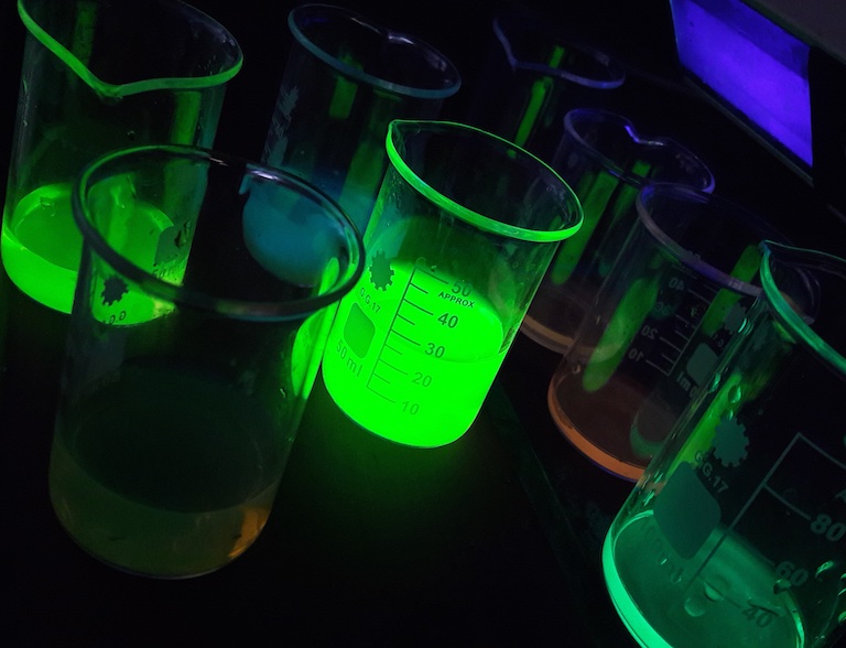 Bild aus halber Höhe in einem halbdunklen Labor auf Bechergläser, deren Flüssigkeiten neongrün leuchten.